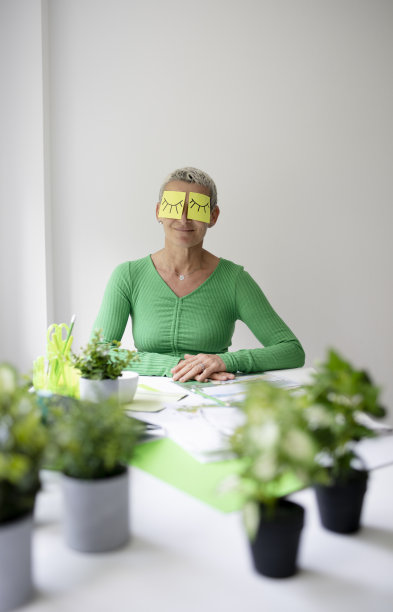 短发的中年妇女坐在摆满绿色植物和工作笔记的办公桌前，脸上画着眼睛的便签纸图片下载