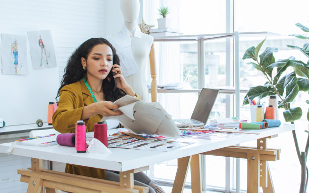 专业女裁缝穿着现代西装，工作，使用手机，在家工作室与客户交谈，制作、设计时装、服装。小生意，生活方式的概念。图片下载