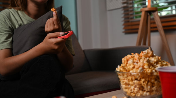 在家看电视，一边吃爆米花，一边拿着遥控器的年轻女子。休闲活动，放松，爱好概念。图片下载