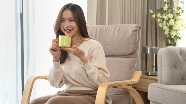 幸福的女人穿着温暖的毛衣喝热茶，在一个明亮的冬天的早晨在家里的扶手椅上放松。图片下载