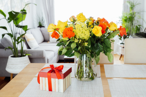 黄色和橙色的玫瑰在花瓶和礼品盒红丝带在厨房柜台桌上与开放空间客厅背景。感恩节、生日、母亲节或情人节的礼物。图片下载
