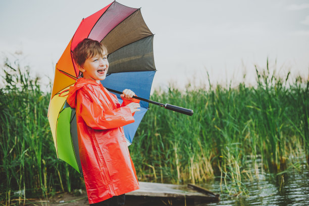 公园里一个带着彩虹伞微笑的男孩的肖像。孩子肩上撑着五颜六色的伞。欢快的孩子，穿着红色雨衣，在户外撑着五颜六色的伞图片下载