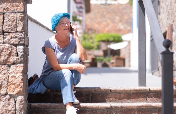 微笑迷人的高级妇女与蓝色帽子坐在户外楼梯上使用手机。在阳光下享受假期或退休生活的老年妇女。图片下载