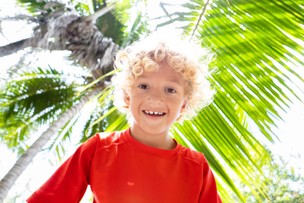 沙滩的乐趣。棕榈树下的卷毛男孩。图片下载