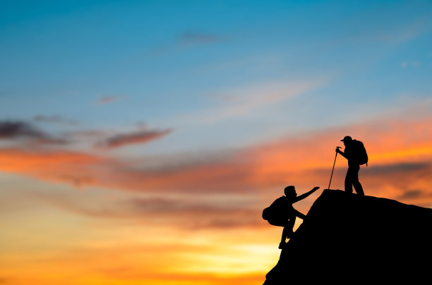 第二个背双肩包的剪影在日出时爬上悬崖。他费了很大的劲才爬到山顶。他们互相帮助到达山顶。图片下载