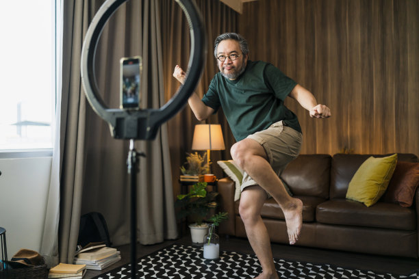 快乐的男性视频记录舞蹈在社交媒体上播出，男人在家里的客厅跳舞，有趣的庆祝有趣的病毒舞蹈自由周末。一名亚洲男子试图在智能手机上为视频直播练习舞蹈图片下载