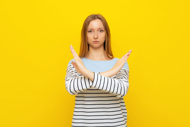 拒绝。年轻严肃的女人与金发显示停止手势交叉的手，穿着休闲条纹运动衫，站在黄色的背景图片下载