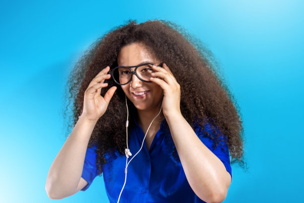 美丽的非洲裔美国女孩戴上眼镜，用耳机听古典音乐。孤立在蓝色背景上图片下载