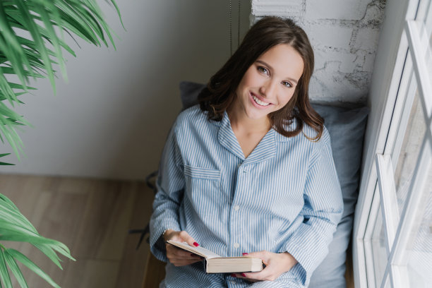 照片上开朗的女人读着浪漫的故事，拿着书，穿着休闲的衣服，在舒适的房间的窗台上摆姿势。从以上观点。满意的女模特心情好，看教科书。图片下载