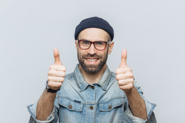 快乐的大胡子男模特戴着眼镜，戴着时髦的帽子，穿着牛仔夹克，一直竖起大拇指，对某事表示赞同和同意，有着愉快的表情，孤立在灰色的墙上图片下载