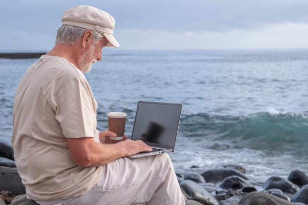 一位活跃的老人坐在沙滩上用笔记本电脑。年长的大胡子男性拿着咖啡杯在海上欣赏日落，地平线在水面上图片下载