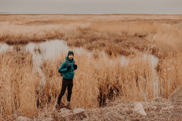 照片中活跃的男性穿着时髦的衣服，站在靠近小湖和黄色秋天的田野的石头上，背着双肩包，复制空间为您的广告内容或信息图片下载