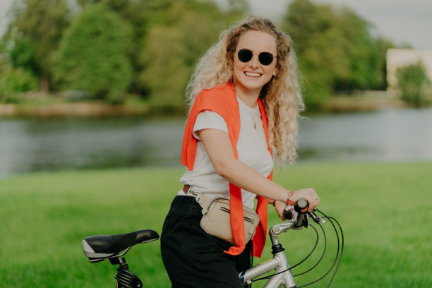 漂亮的女骑自行车的人停在美丽的河边，戴着墨镜，白t恤，肩上套头衫，和她的交通工具在户外摆姿势，表达积极的情绪，享受美妙的旅行图片下载