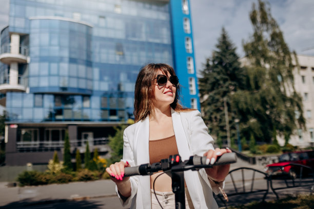 美丽的年轻女子戴着太阳镜和白色西装站在她的电动摩托车附近的现代建筑和看向别处图片下载