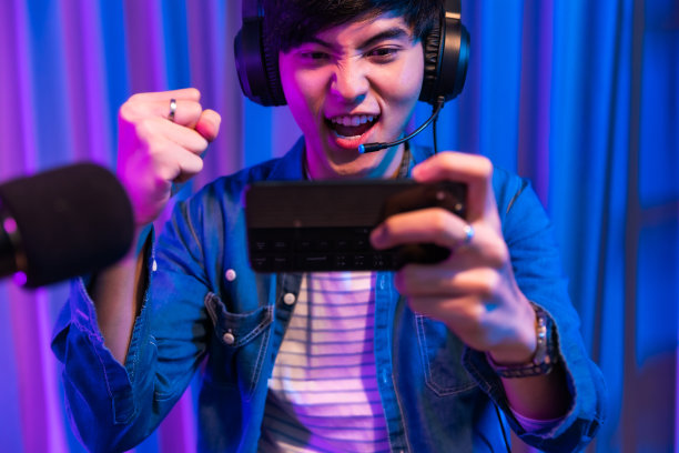 用智能手机玩游戏。年轻的亚洲帅哥坐在椅子上，手里拿着手机。霓虹灯室戴着耳机在线手机游戏退出流媒体。在线电竞流媒体游戏，家庭隔离。活动。图片下载