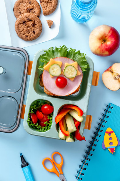 为孩子们准备的学校饭盒里的食物以滑稽的面孔的形式出现。学校午餐盒，桌上有三明治、蔬菜、水和文具。健康饮食习惯观念。回归学校理念图片下载