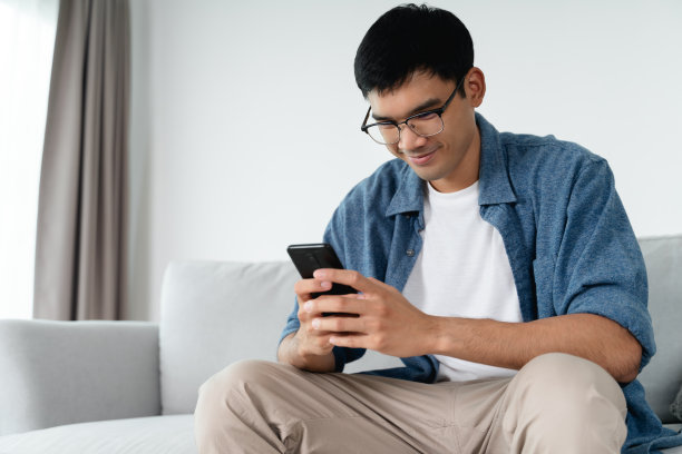 幸福的亚洲男人在家里客厅的沙发上使用智能手机，搜索数据和社交媒体。图片下载