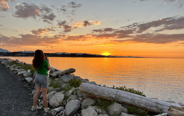 一个十几岁的女孩在一件绿色的t恤拍摄太平洋上美丽的日落，她手里拿着手机，金色的红色头发，可见的石头日落，大海和天空图片下载