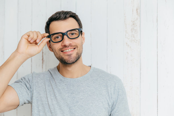 留着胡茬的迷人男模，戴着眼镜看起来很开心，和别人愉快地交谈，穿着休闲t恤，在白色木制背景下摆姿势。生活方式的概念。图片下载