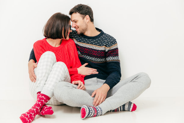 照片中，一对相爱的情侣盘腿坐在白色的地板上，深情地望着对方，拥抱着，享受着在一起的感觉，孤立在白色的背景上。人，爱与爱人的观念图片下载