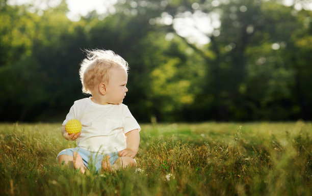 一个可爱的婴儿坐在公园的空地上，手里拿着一个玩具，向右看图片下载