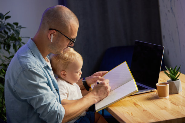 年轻的父亲抱着年幼的儿子在笔记本电脑上远程工作图片下载
