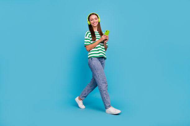 全身照片酷年轻女士走听音乐戴耳机t恤牛仔裤靴子孤立的蓝色背景图片下载