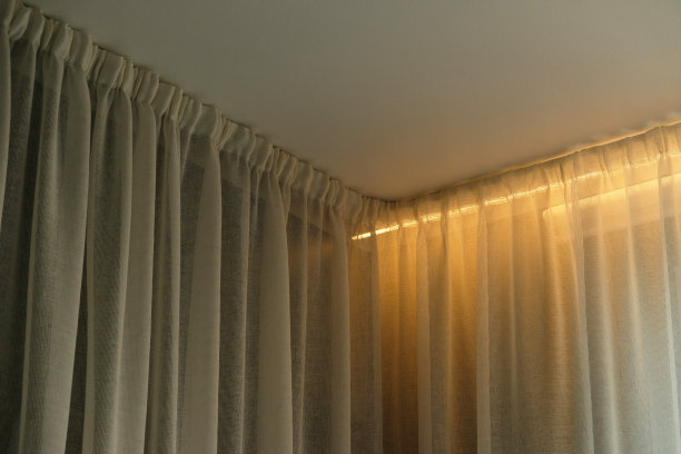 有窗帘和白色窗帘或薄纱的角状飞檐。白色的墙壁，天花板，飞檐壁龛，窗户上的窗帘。墙角藏帘杆。天花板下窗帘后面的照明。图片下载
