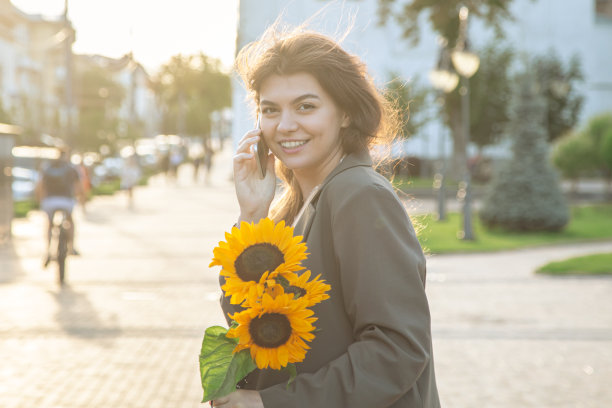 日落时分，一位手持一束向日葵的年轻女子正在讲电话。图片下载