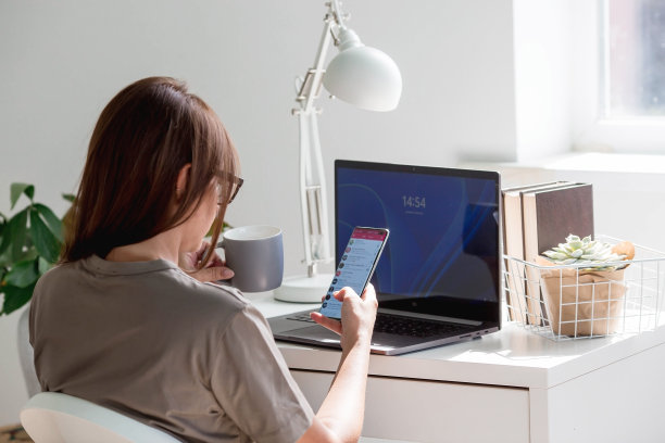 一个年轻，快乐，美丽的自由职业女性坐在家里的办公室里，手里拿着笔记本电脑和智能手机。女性在家工作或在网上商店购物。图片下载