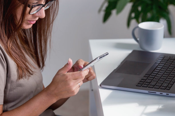 一个年轻，快乐，美丽的自由职业女性坐在家里的办公室里，手里拿着笔记本电脑和智能手机。女性在家工作或在网上商店购物。图片下载