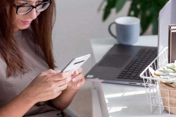 一个年轻，快乐，美丽的自由职业女性坐在家里的办公室里，手里拿着笔记本电脑和智能手机。女性在家工作或在网上商店购物。图片下载