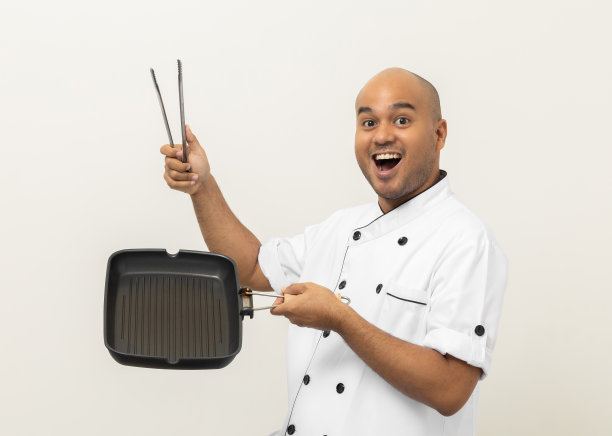 年轻的亚洲男性厨师穿着制服，用铁制烤盘和钳子烹饪厨房用具。厨师职业厨师在厨房、餐厅和酒店工作。图片下载