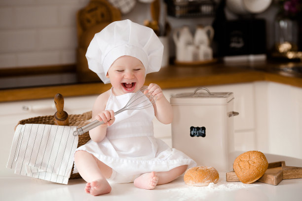 孩子们的厨师。漂亮的小男孩，围着白围裙，戴着厨师帽，手里拿着搅拌器。背景是厨房的内部。烹饪概念。图片下载