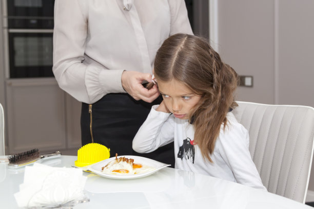 近距离的照片，快乐的女孩和她的长发坐在早餐桌旁，她的母亲在家里梳头发，他们穿着白色的衬衫，人们的生活方式的概念图片下载