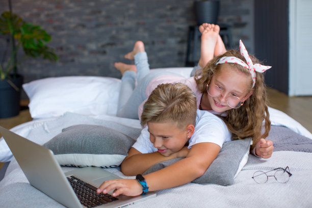金发孩子——一个男孩和一个女孩躺在床上，在笔记本电脑前学习和玩耍图片下载