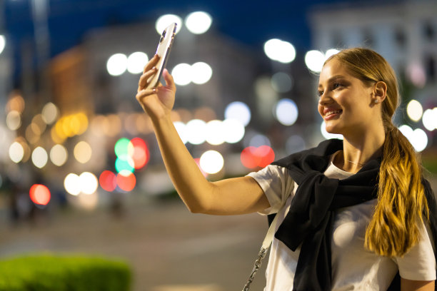 年轻女子使用智能手机在夜市背景散景灯，女子自拍，在线wi-fi互联网图片下载