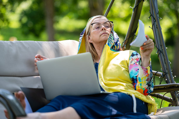 高级妇女与笔记本电脑和文件工作在花园摇椅，绿色家庭办公室的概念图片下载