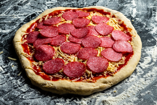 黑乎乎的桌子上撒着面粉，放着意大利辣香肠的生披萨。图片下载