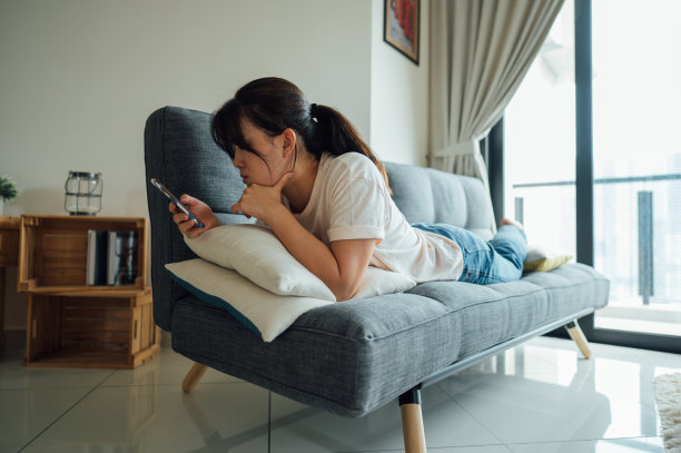 漂亮年轻的亚洲女人，在她的客厅沙发上放松，同时使用她的智能手机的社交媒体和上网。图片下载