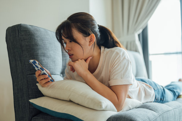 一名亚洲女性躺在客厅的沙发上用智能手机社交媒体图片下载