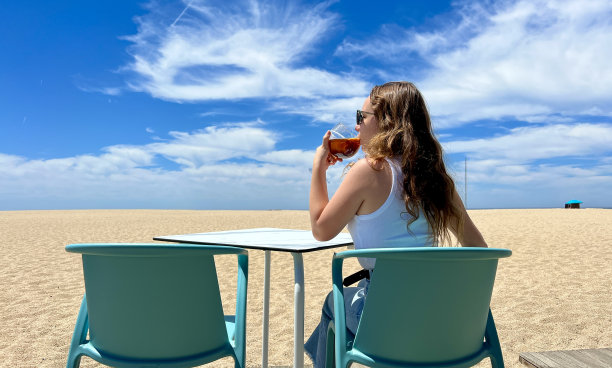 一个十几岁的女孩坐在海洋或海的背景在她的手，她有一杯冰茶，她穿着白色的t恤，她的头发是宽松的，她的背影对着相机图片下载