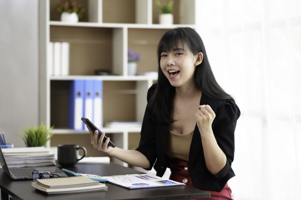 快乐的年轻亚洲商业女性成功兴奋地举起手在办公室与智能手机欣喜。图片下载