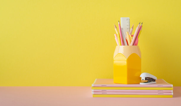 学校附件的概念。文具的照片在粉红色的桌面铅笔盒和迷你订书机在黄色的墙壁背景与copyspace的记事本堆栈图片下载