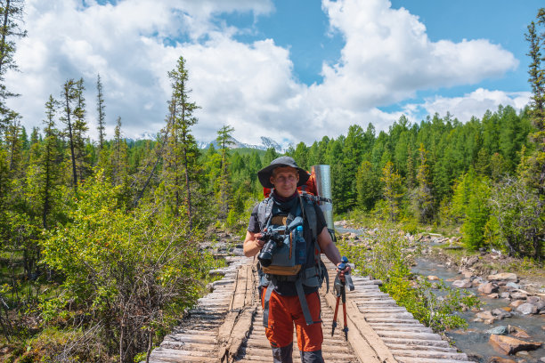 在明亮的阳光下，背着大背包和相机的旅行者在森林里的木桥上穿过山河。阳光明媚的日子里，徒步穿越针叶林，穿越河流，风景生动。图片下载