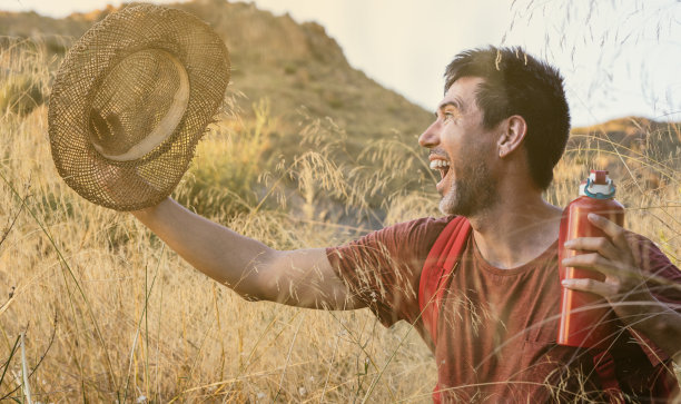 年轻的成年白人男子笑在幸福的帽子和红色背包。他在一个沙漠地区，在日落的黄金时间旅行。在大自然中自由和冒险的概念。图片下载
