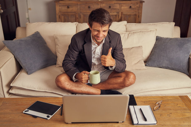 穿着西装和短裤的年轻人，一边喝着咖啡，一边在家里的沙发上用笔记本电脑进行视频通话。他用手做了一个喜欢的手势。图片下载