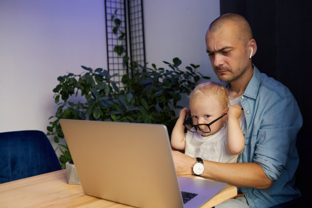 年轻的父亲抱着年幼的儿子在笔记本电脑上远程工作图片下载