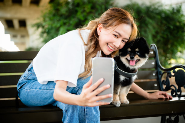 快乐的年轻亚洲女人坐在长椅上和照片自拍在公园里和她的狗。宠物情人的概念图片下载