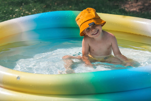 一个炎热的夏日。孩子在游泳池里游泳，溅起水花。一个戴着黄色巴拿马草帽的快乐男孩对着镜头微笑。水溅向四面八方。和孩子们一起过暑假的概念。图片下载